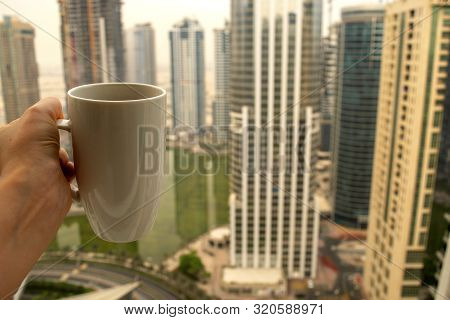 Female Hand Holds Big White Cup Of Morning Hot Drink - Coffee Or Tea With City Skyscrapers View Of M