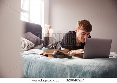 Male College Student Lies On Bed In Shared House Working On Laptop