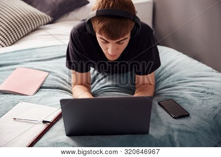 Male College Student Wearing Headphones Lies On Bed In Shared House Working On Laptop