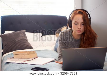 Female College Student Wearing Headphones Lies On Bed In Shared House With Mobile Phone Using Laptop