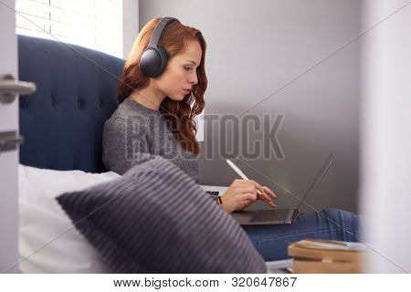Female College Student Wearing Wireless Headphones Sitting On Bed In Shared House Using Laptop