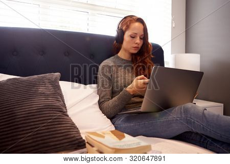 Female College Student Wearing Headphones Works On Bed In Shared House With Laptop And Mobile Phone