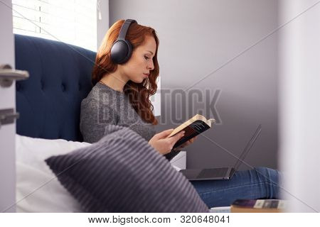 Female College Student Wearing Wireless Headphones Sitting On Bed In Shared House Using Laptop