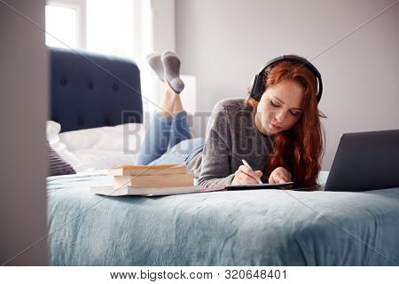 Female College Student Wearing Headphones Lies On Bed In Shared House With Mobile Phone Using Laptop