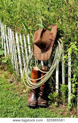 Cowboy Hat And Lasso Hanging On A Short White Picket Fence Over A Well Worn Pair Of Cowboy Boots.