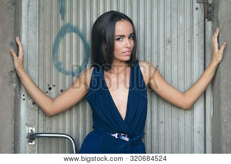 Portrait Of A Fashionable Woman In A Blue Dress With A Neckline. Industrial Wall In The Background