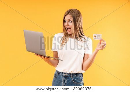 Image of astonished blond woman xpressing wonder while holding laptop computer and credit card isolated over yellow background in studio