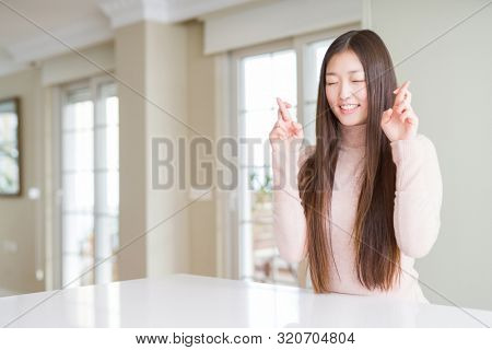 Beautiful Asian woman wearing casual sweater on white table smiling crossing fingers with hope and eyes closed. Luck and superstitious concept.