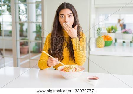 Young woman eating a bowl of Asian rice using chopsticks cover mouth with hand shocked with shame for mistake, expression of fear, scared in silence, secret concept