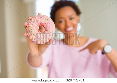 Young african american woman eating pink sugar donut with surprise face pointing finger to himself