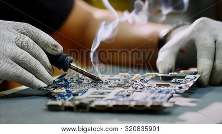 Repairman In Gloves Is Soldering Motherboard Of Computer Device In Workshop.