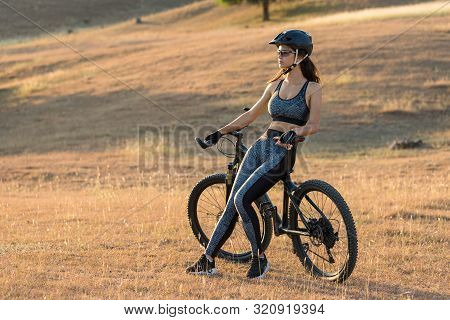 Girl On A Mountain Bike On Offroad, Beautiful Portrait Of A Cyclist At Sunset, Fitness Girl Rides A 