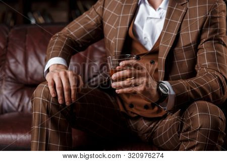 Young Successful Man In A Brown Kosmtyum Drinks Whiskey. Bearded Businessman Sitting In A Library On