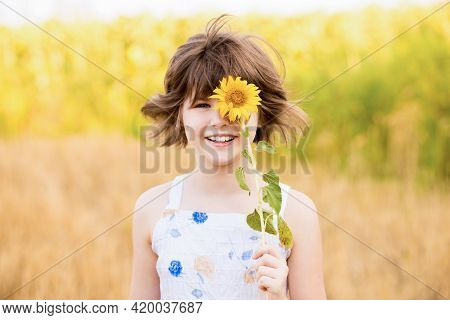Cute Child Girl Wear Dress With Sunflower In Summer Field. Happy Little Girl Hide Eye With Sunflower