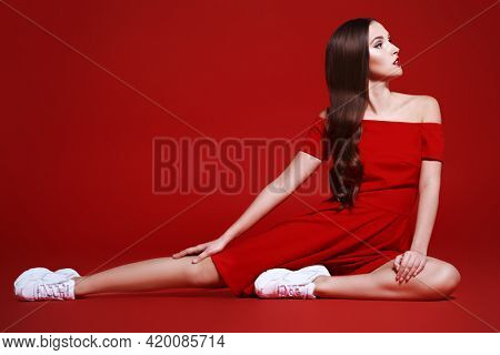 Beautiful brunette girl posing at studio in red dress and white sneakers. Red background. Sport chic style.