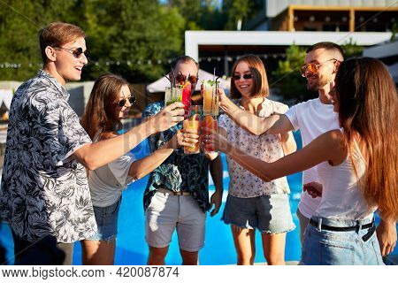 Group Of Friends Having Fun At Poolside Summer Party, Clinking Glasses With Colorful Summer Cocktail