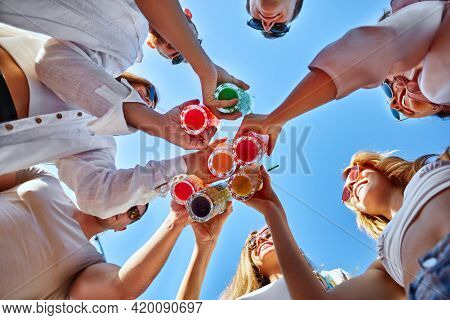 Low Angle View Of Friends Having Fun At Poolside Summer Party, Clinking Glasses With Colorful Summer