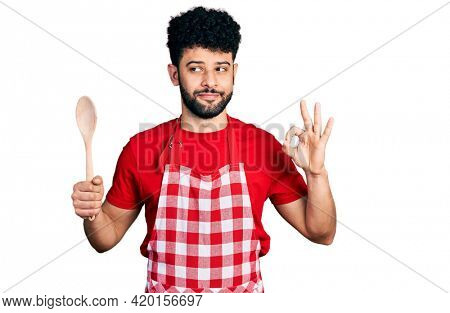 Young arab man with beard wearing baker uniform holding wooden spoon smiling looking to the side and staring away thinking. 