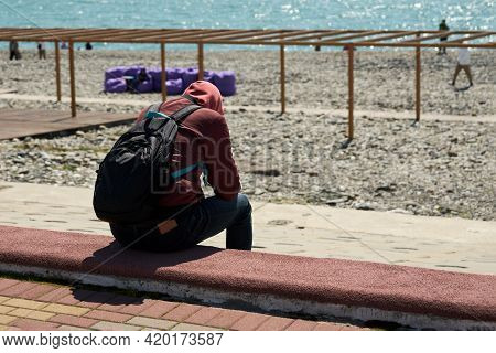 A Lone Figure Of A Young Man With A Backpack On His Back, Sitting On The Sparsely Populated Embankme