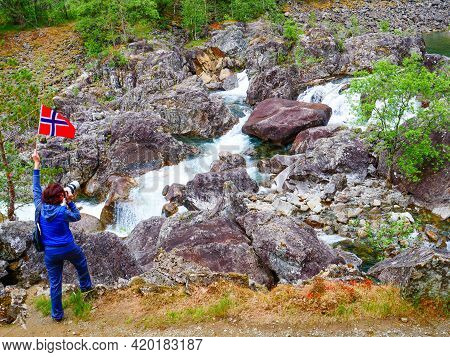 Tourist Woman With Norwegian Flag And Camera Walking Trough Mountains Path In Allmannajuvet Area Sau