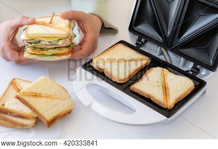 Hands Hold Cooked Fresh Sandwich With Cheese, Bacon And Vegetable Near Toster On The White Table.