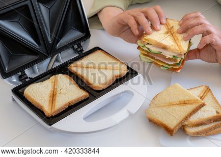 Hands Hold Cooked Fresh Sandwich With Cheese, Bacon And Vegetable Near Toster On The White Table.