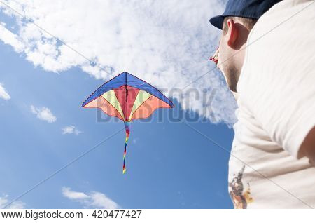 Young Man Holding Colorful Bright Rainbow Kite Hovering In The Blue Sky With White Clouds