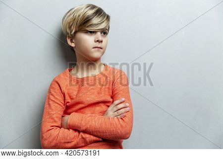 A Serious Boy Of 9 Years Old In An Orange Jacket Stands Against The Wall. The Arms Are Crossed Over 