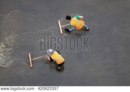Two Workers Cleans The Street After The Rain. Janitors With Brushes Washes Wet City Road, View From 