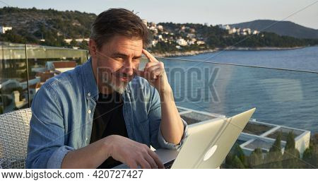Happy man working online with laptop computer on terrace. Home office, browsing internet, tele working, video chat. Businessman on vacation at sea. Escape, freedom, getaway.