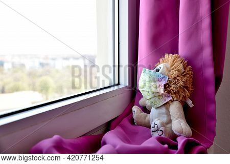 A Teddy Bear Waering A Medical Protective Mask Sits On A Windowsill And Looks Out The Window. Sadnes