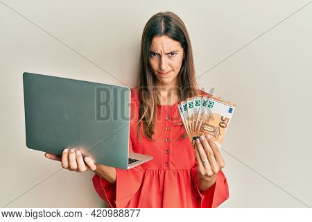 Young beautiful woman holding laptop and 50 euros skeptic and nervous, frowning upset because of problem. negative person. 