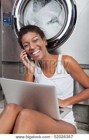 Portrait of happy young African American woman with laptop using mobilephone in laundry