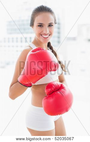 Smiling sporty brunette wearing red boxing gloves in bright room