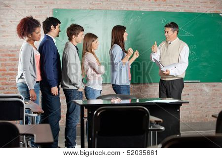 Teacher gesturing thumbsup sign to successful female student with classmates standing in row at classroom