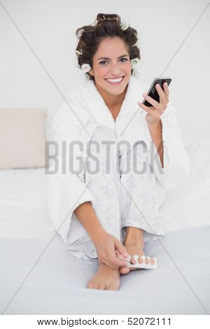 Happy smiling brunette applying nail polish to toes and texting in bedroom