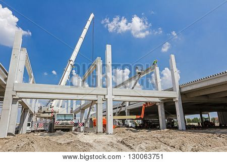 Mobile crane is operating and worker is assembly concrete joist in high place. Height worker is placing truss on building skeleton.