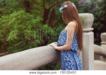 A young girl stands on a bridge on a background of green trees and looks down. The girl turned sideways.