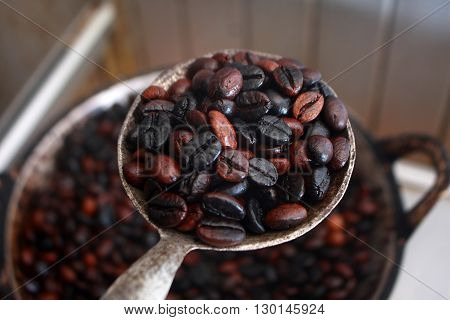 Dark roasted coffee beans in scoop with blurred fresh roasted coffee beans in pan on background
