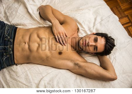 Shirtless sexy male model lying alone on his bed in his bedroom, looking at camera with a seductive attitude