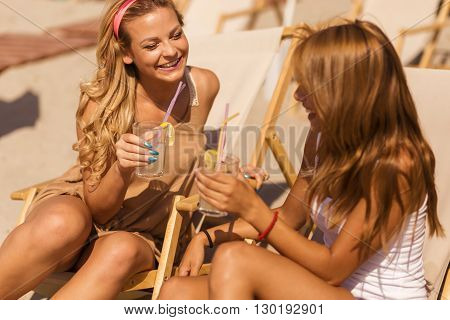 Two beautiful young girls lying on a sunbed on the beach cheering with a lemonade and chatting