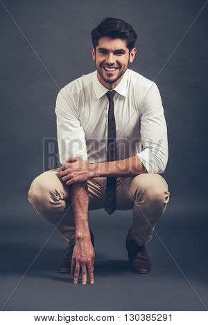 Can you resist his smile? Full length of confident young handsome man looking at camera with smile while sitting crouched against grey background