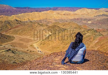 The girl relaxes after the hiking sitting on peak of Zefahot Mount and watch the amazing colorful landscapes Eilat Israel.