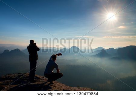 Two Friends. Hiker Thinking And Photo Enthusiast Takes Photos  Stay On Cliff. Dreamy Fogy Landscape,
