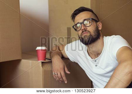 Young man moving in a new appartment sitting on the floor drinking take away coffee surrounded with cardboard boxes