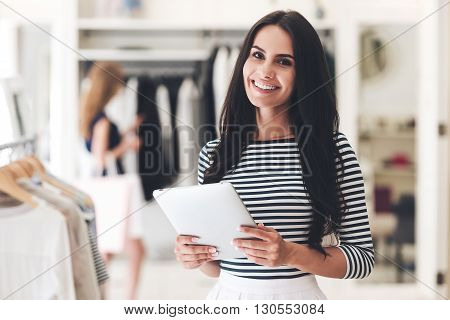Starting new business. Beautiful young woman holding digital tablet and looking at camera with smile while standing at the clothing store
