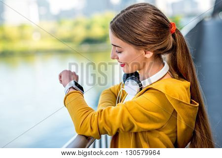 Young sport woman in yellow sweater looking at smart watch on the modern bridge with skyscrapers on the background. Morning exercise in megacity