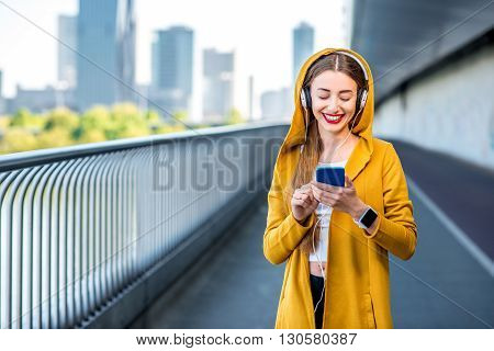 Young sport woman in yellow sweater listening to the music with smart phone on the modern bridge with skyscrapers on the background. Morning exercise in megacity