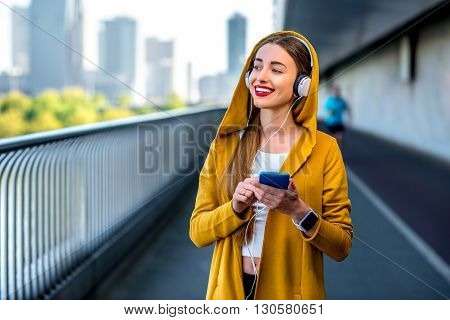 Young sport woman in yellow sweater listening to the music with smart phone on the modern bridge with skyscrapers on the background. Morning exercise in megacity
