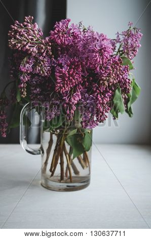 Still life with blooming spring branches of lilac flowers in vase on dark background. Home decoration in a rustic style. Violet bouquet on white windowsill in room.